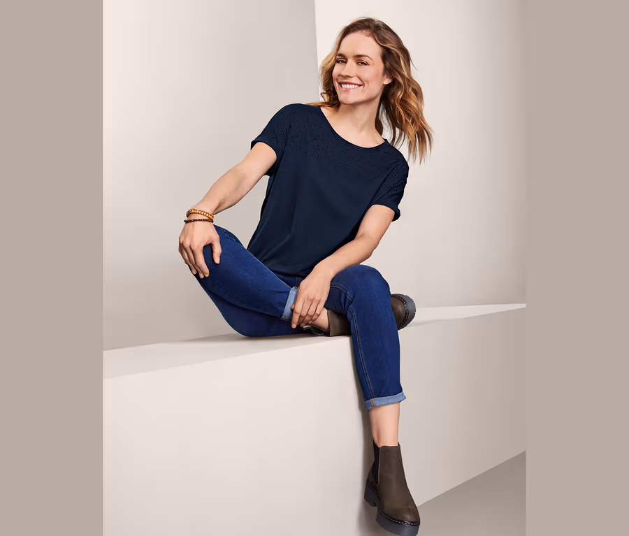 Frau sitzt und trägt blaue Jeans und ein Kurzarmshirt mit Stickerei.