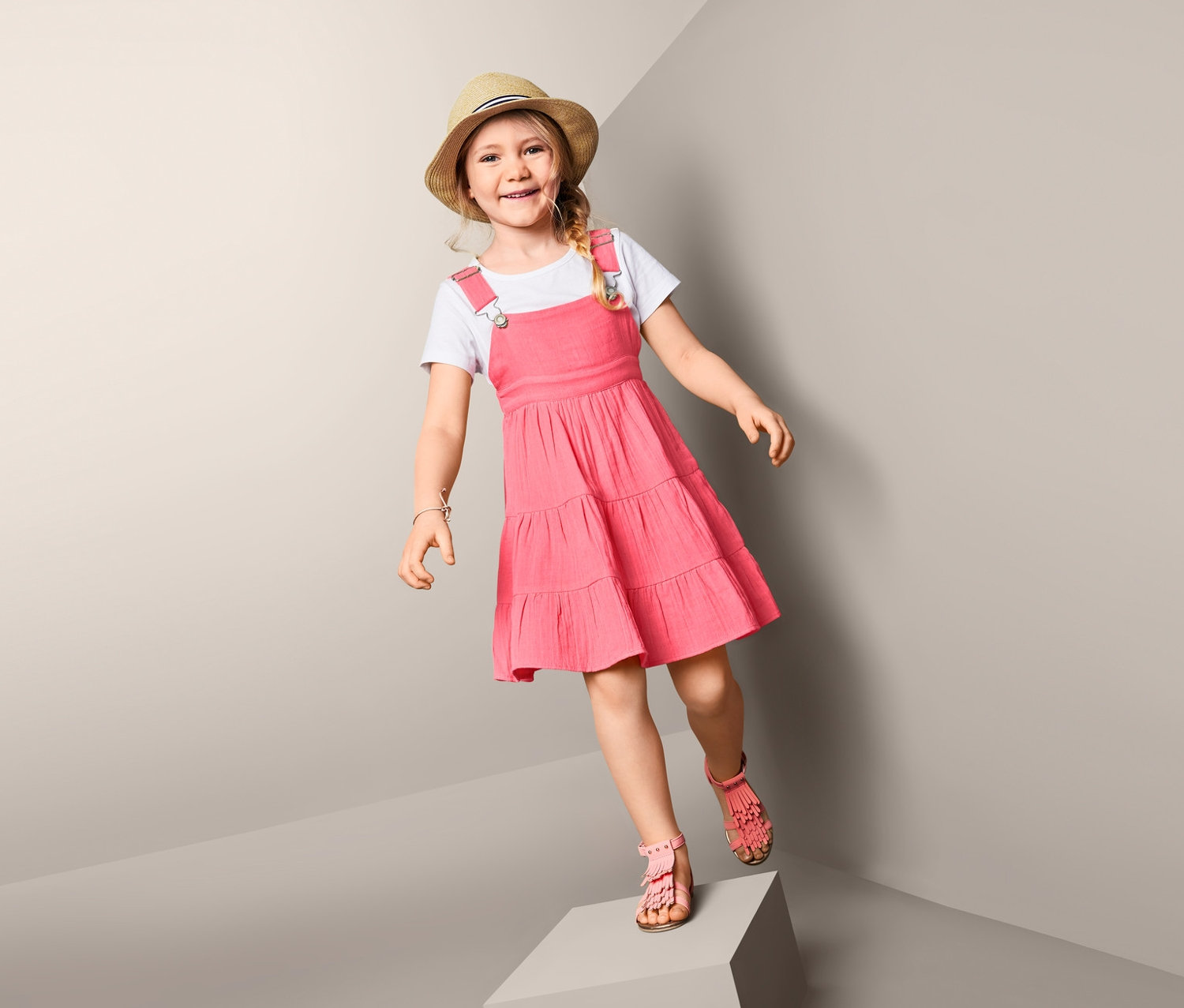 Ein Mädchen steht auf einem Würfel und trägt ein Trägerkleid und einen Hut.