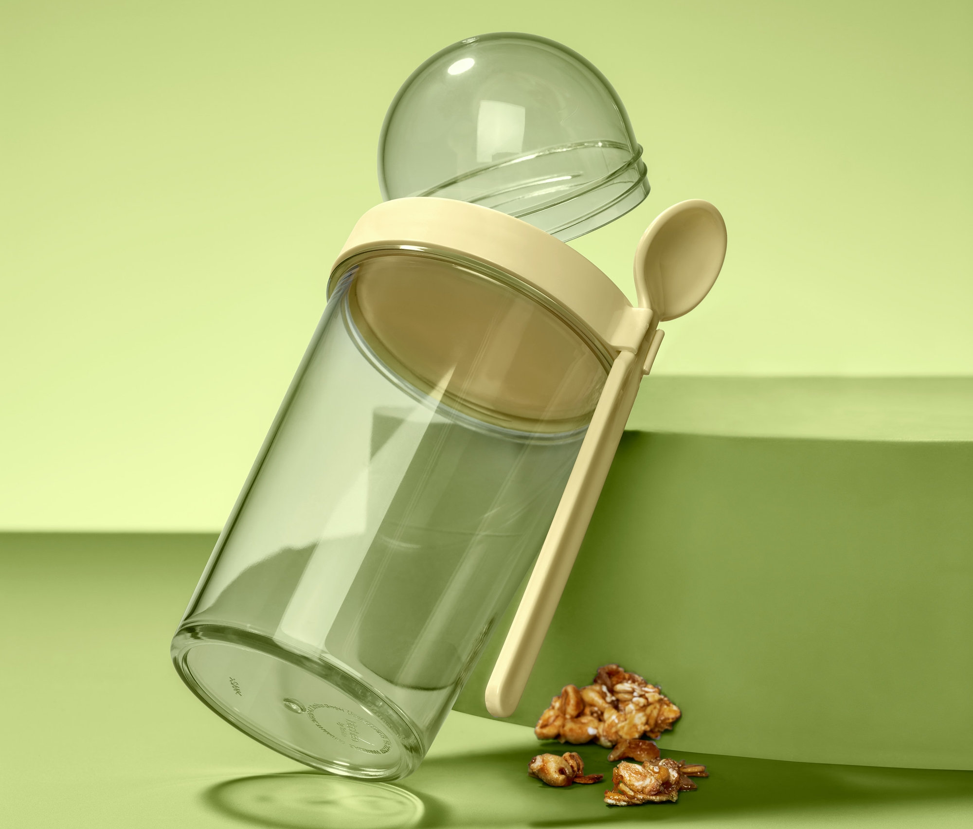 Ein durchsichtiger Joghurt-to-go-Becher mit beigem Deckel, Löffel und durchsichtigem Kugelbehälter steht schräg auf einer grünen Oberfläche neben Müslifragmenten.