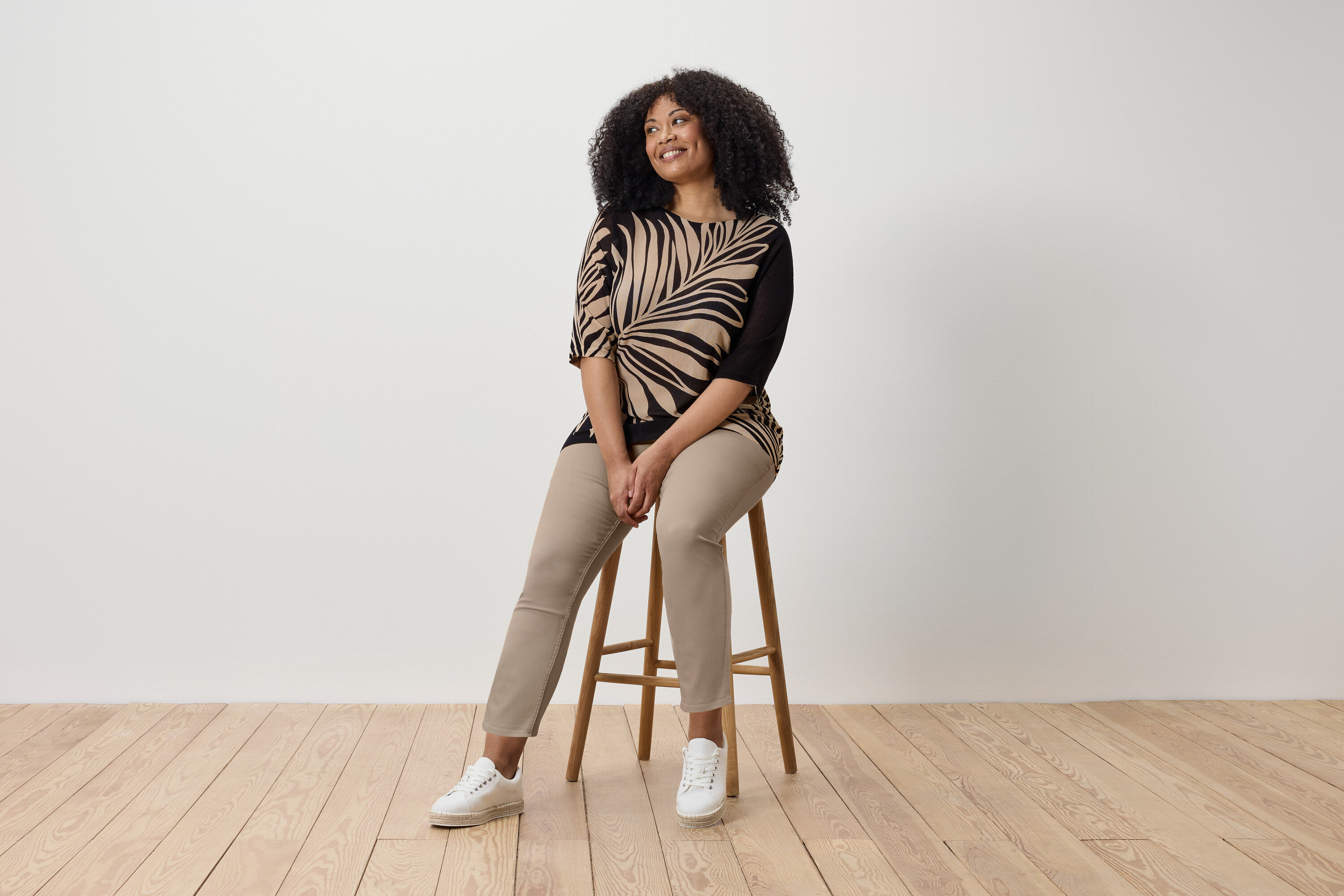 Frau sitzt auf einem Hocker und trägt 7/8-Treggings, ein bedrucktes Oberteil und Turnschuhe.