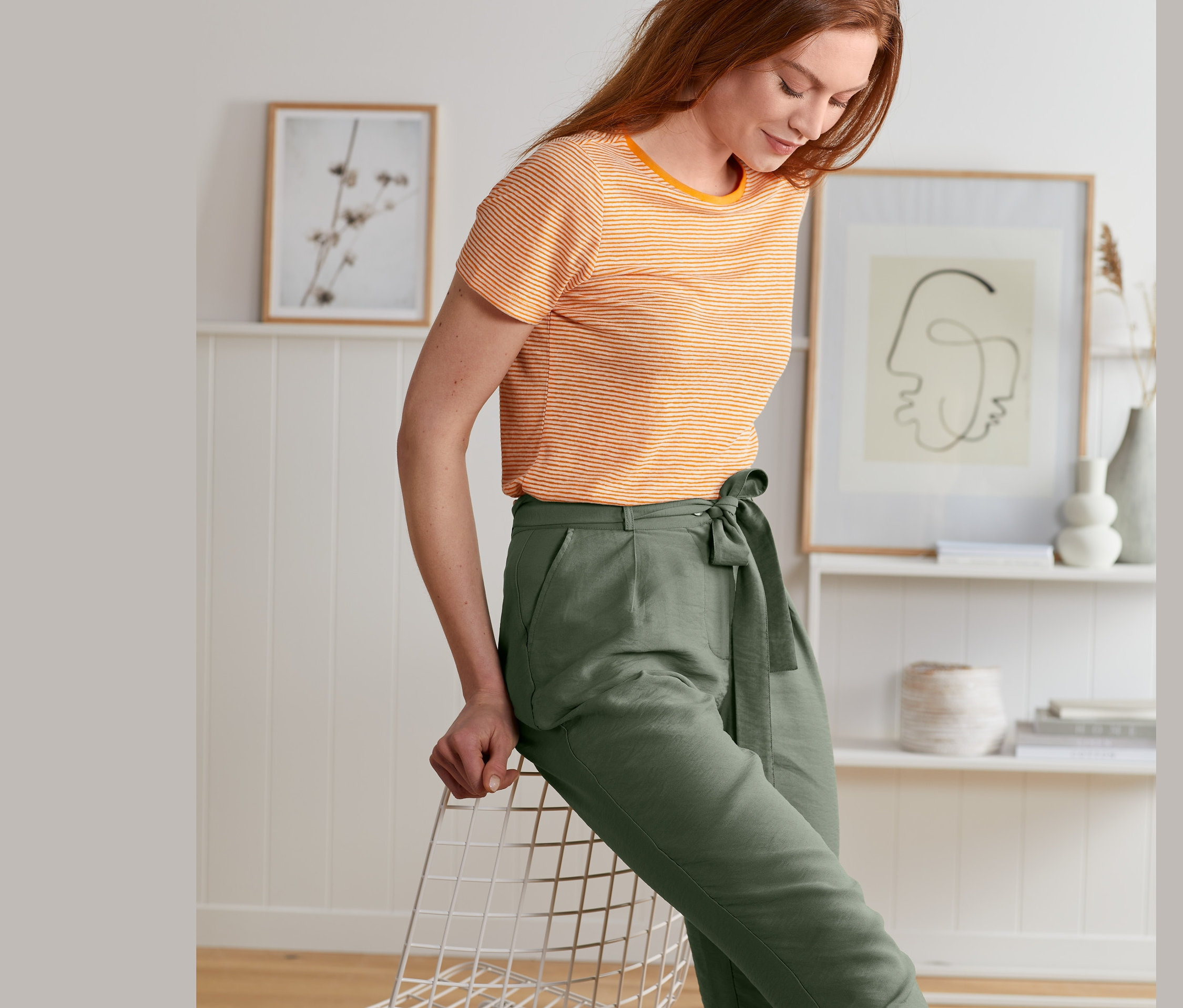 Rothaarige Frau sitzt an einen Stuhl gelehnt und trägt eine olivgrüne Webhose mit Bindegürtel und ein gestreiftes T-Shirt.