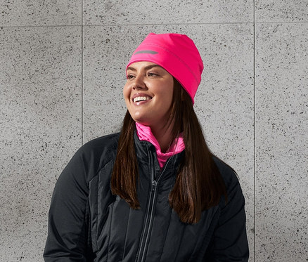 Eine lächelnde Frau sitzt auf einer Bank und trägt eine schwarze Thermojacke, Thermo-Lauftight, eine neonpinke Thermomütze mit Reflektorprint und ein neonpinkes Multifunktionstuch. Sie hält eine Wasserflasche in der Hand.