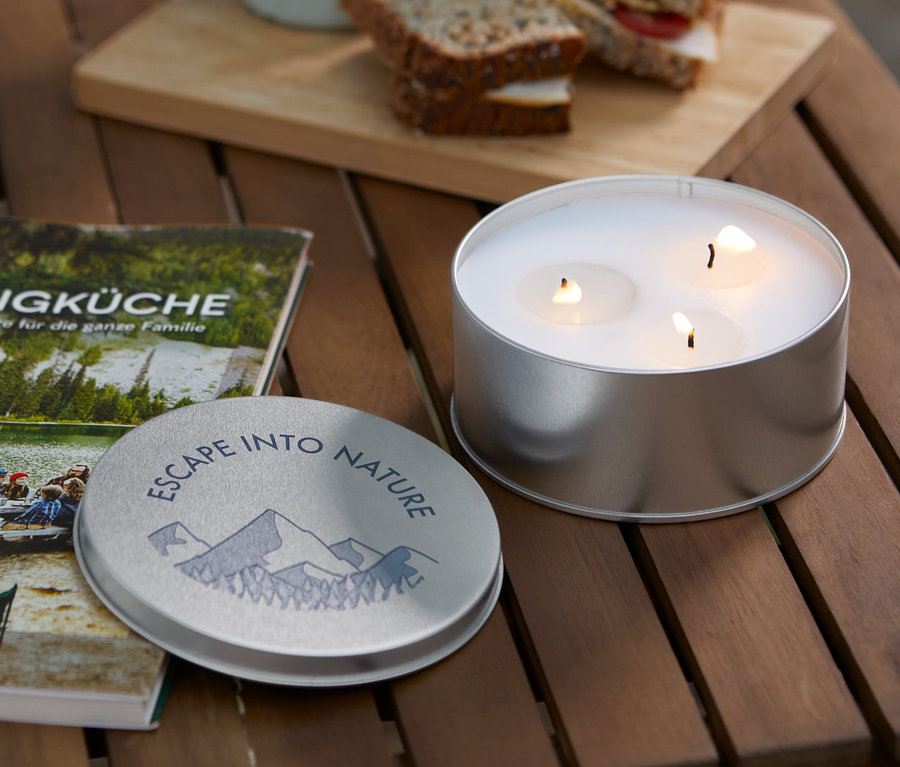 Auf einem Holztisch liegen ein Buch »Campingküche«, eine Citronellakerze, eine Tasse und Sandwiches.
