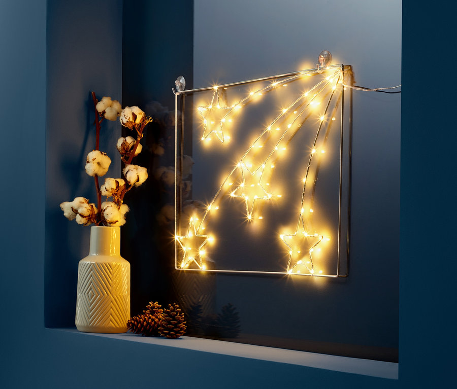 Ein quadratisches LED-Fensterbild mit leuchtenden Sternen neben einer Vase mit Baumwollzweigen und Tannenzapfen.