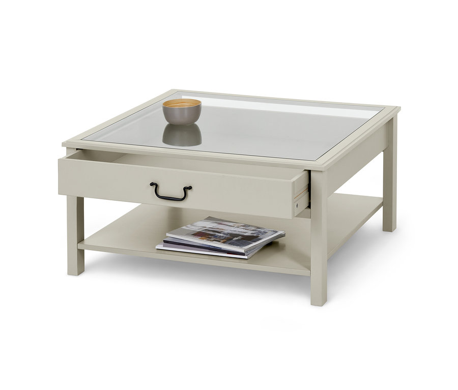 Ein Couchtisch mit Glasplatte, einer offenen Schublade und einer Schale darauf. Darunter befindet sich ein Regal mit Zeitschriften.