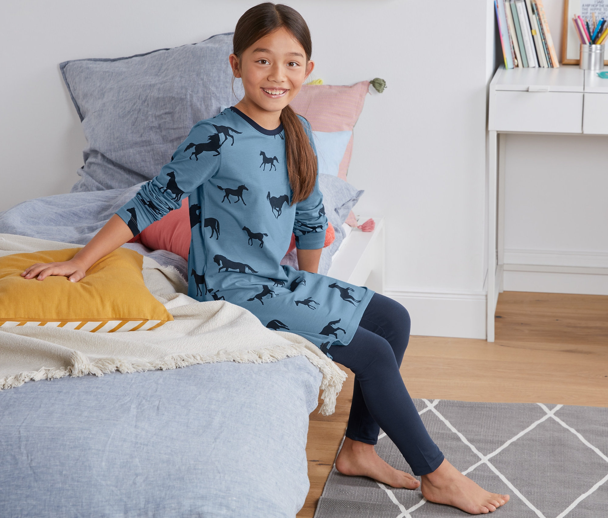 Mädchen sitzt im Jersey-Nachthemd mit Leggings auf dem Bett.