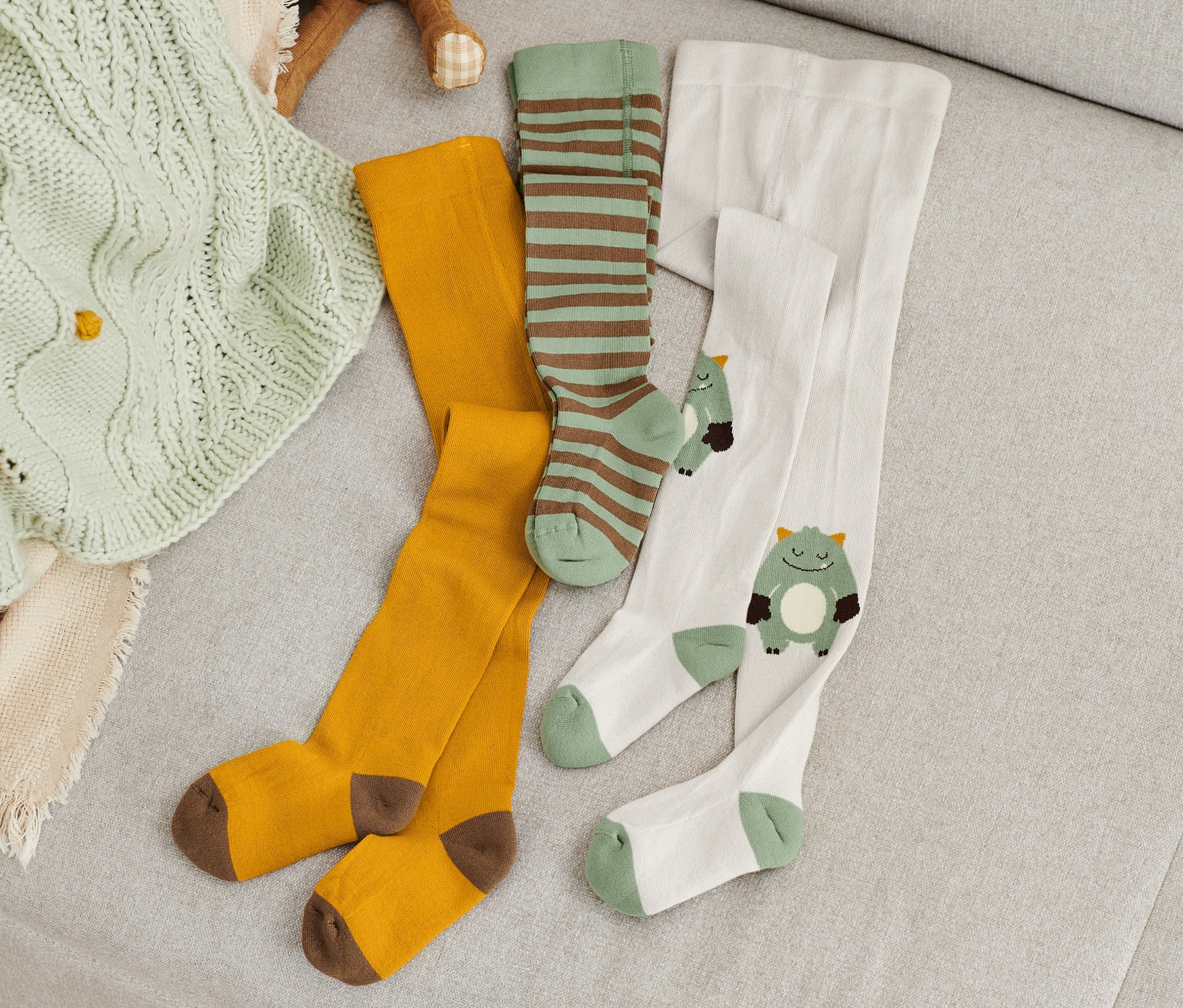 Auf einem Sofa liegen 3 Paar grüne Strumpfhosen neben einem Fuchs-Stofftier und einer Strickdecke.