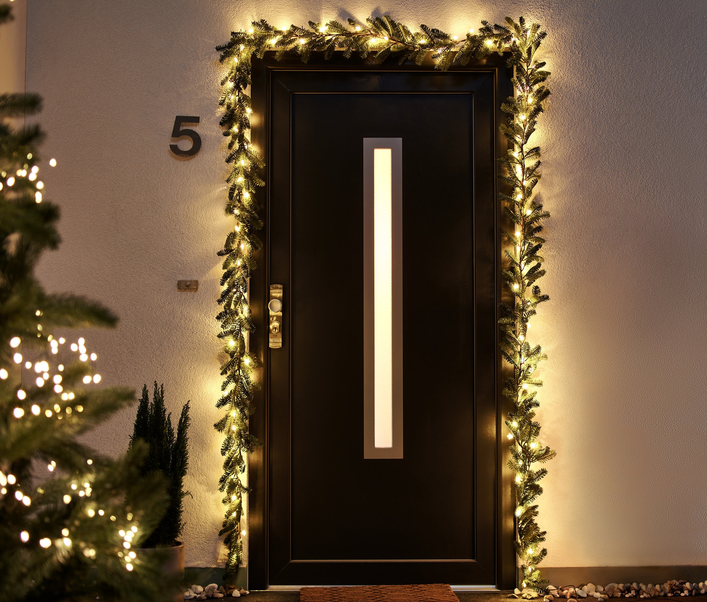 Eine Eingangstür umrahmt von einer LED-Tannengirlande, links ein Weihnachtsbaum mit LED-Lichtern.