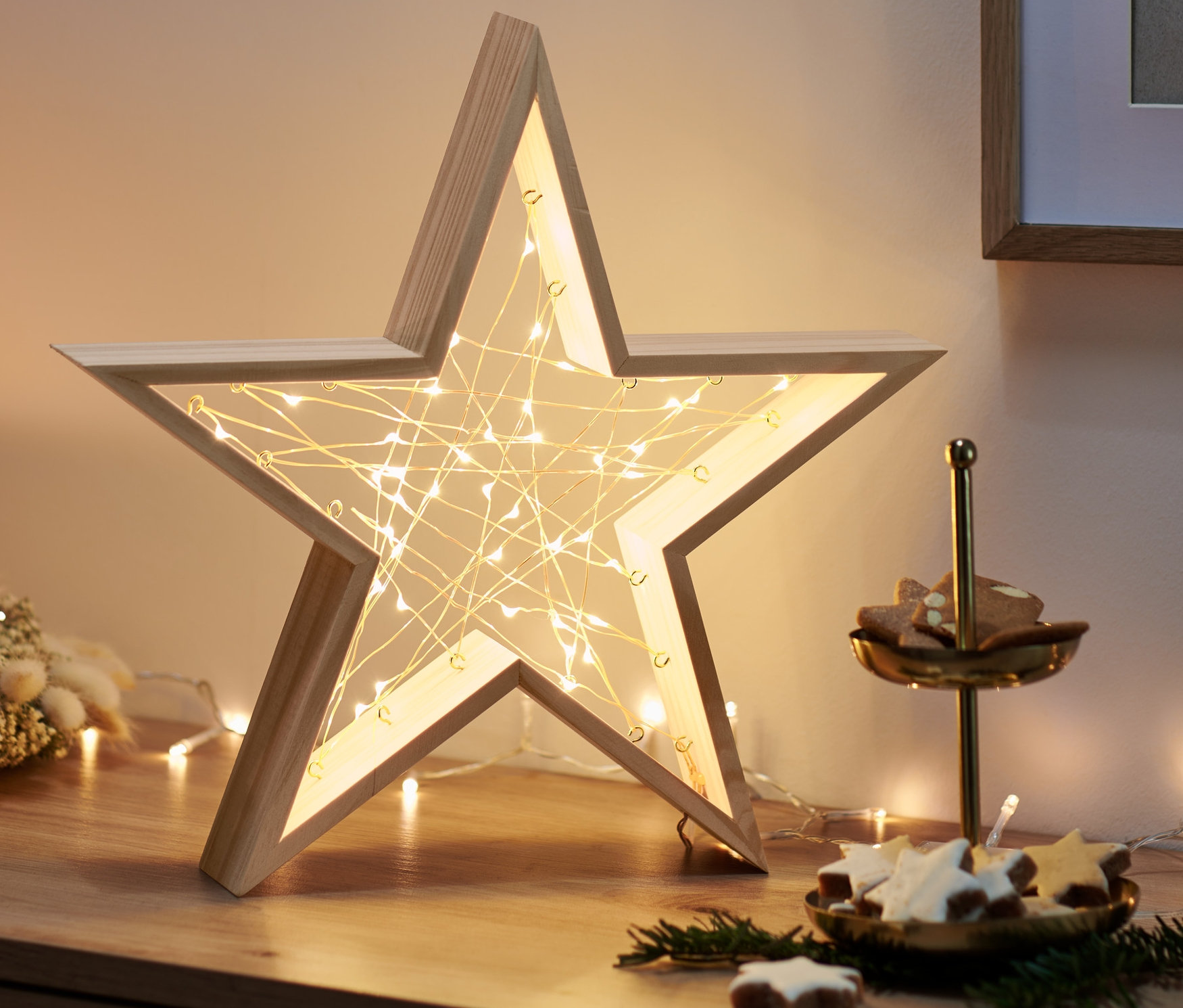 Auf einer Holzkommode stehen ein LED-Holzstern, ein Adventskranz mit Kerze und eine Etagere mit Keksen.