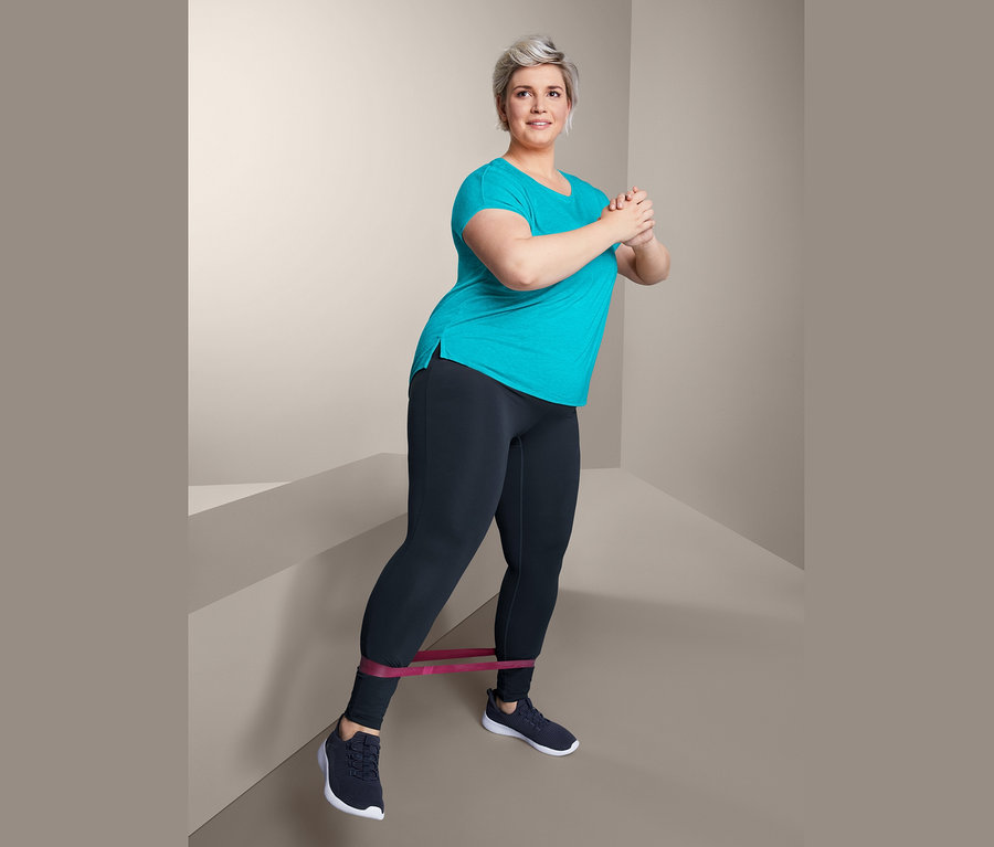 Frau macht Übungen mit einem Band um die Beine, trägt ein Sportshirt und eine Power-Sporttight.