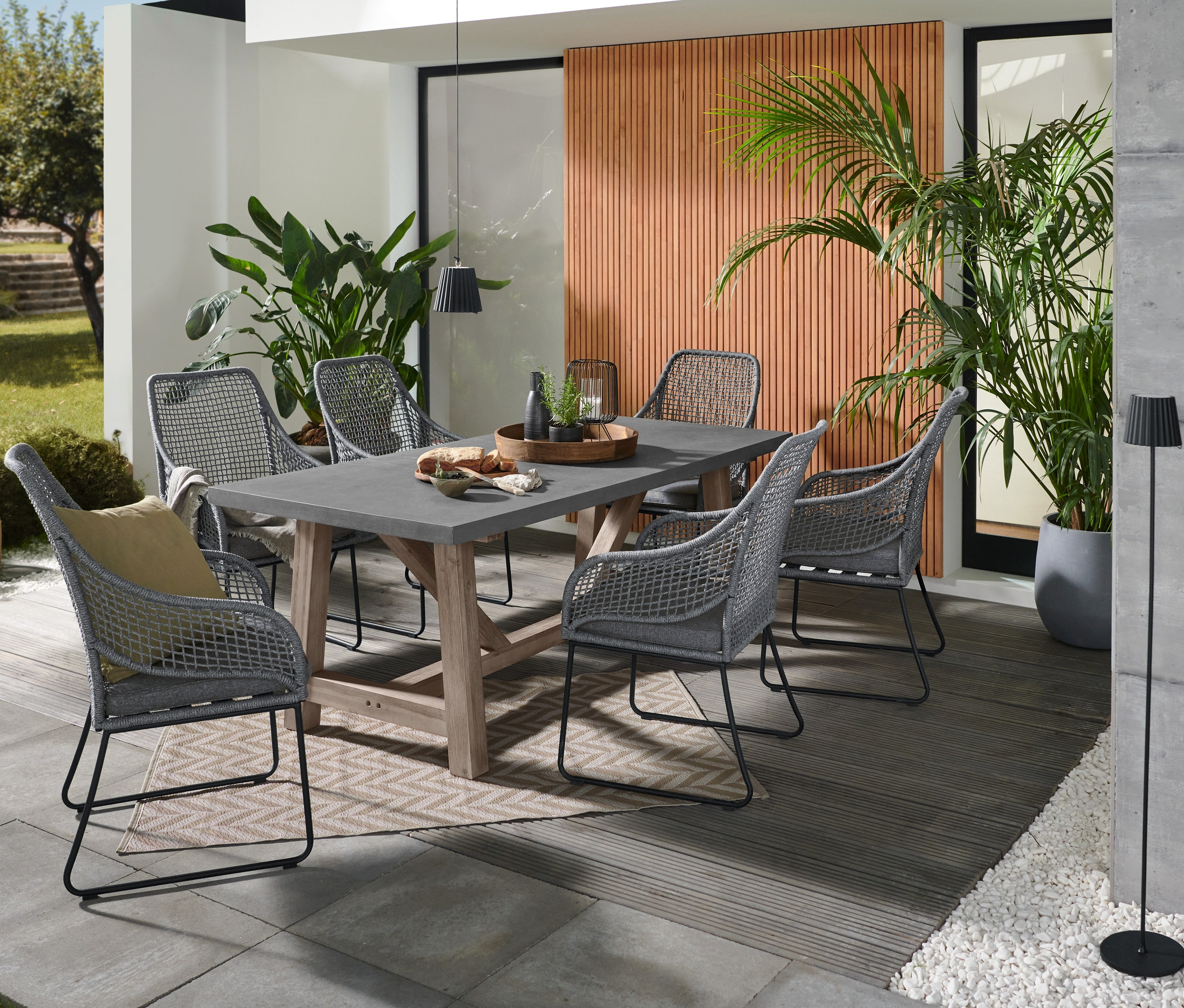 Sechs Hochlehner mit Textilgeflecht stehen um einen Gartentisch mit Beton-Tischplatte auf einem grauen Outdoor-Teppich.