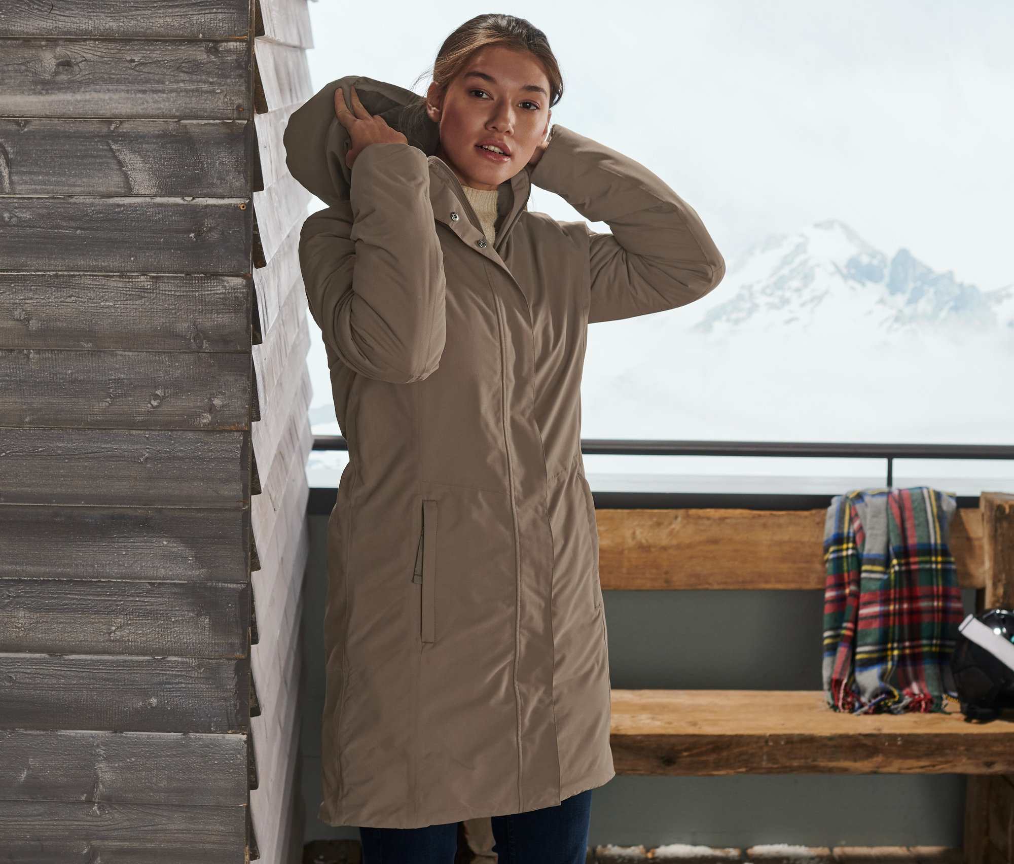 Frau justiert die Kapuze ihres taupefarbenen Thermo-Mantels. Im Hintergrund sind Ski und eine verschneite Landschaft zu sehen.