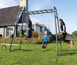 Junge hängt an Ringen des EXIT Outdoor-Fitnessgeräts »MB230«, während Frau an der Klimmzugstange trainiert.