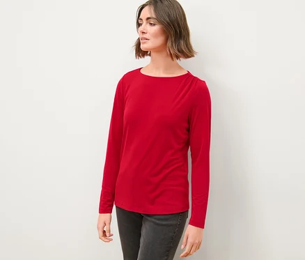 Frau in rotem Blusenshirt mit Raffung und grauen Jeans steht vor einer weißen Wand.