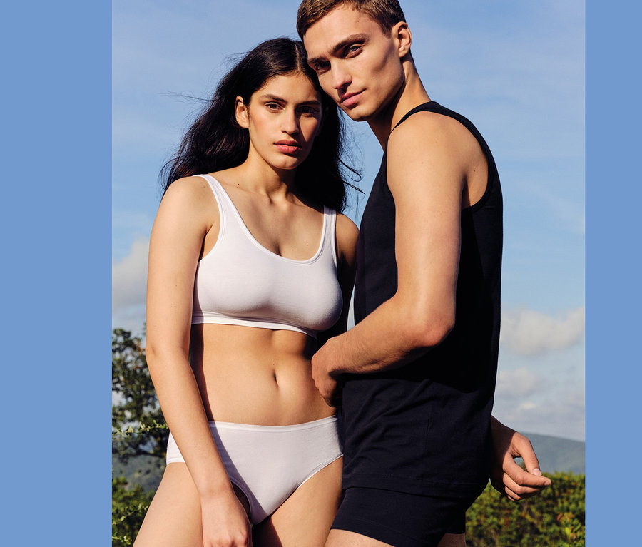 Eine Frau in einem weißen Top und weißen SCHIESSER Hipster (2er Pack) steht neben einem Mann in einem schwarzen Tanktop und schwarzen Boxershorts.