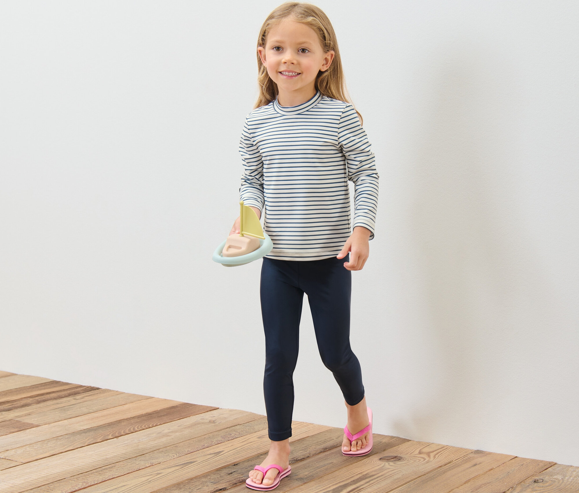 Mädchen hält Spielzeugboot, trägt Kinder-Shirt mit UV-Schutzfaktor 80, Leggings und Zehentrenner.