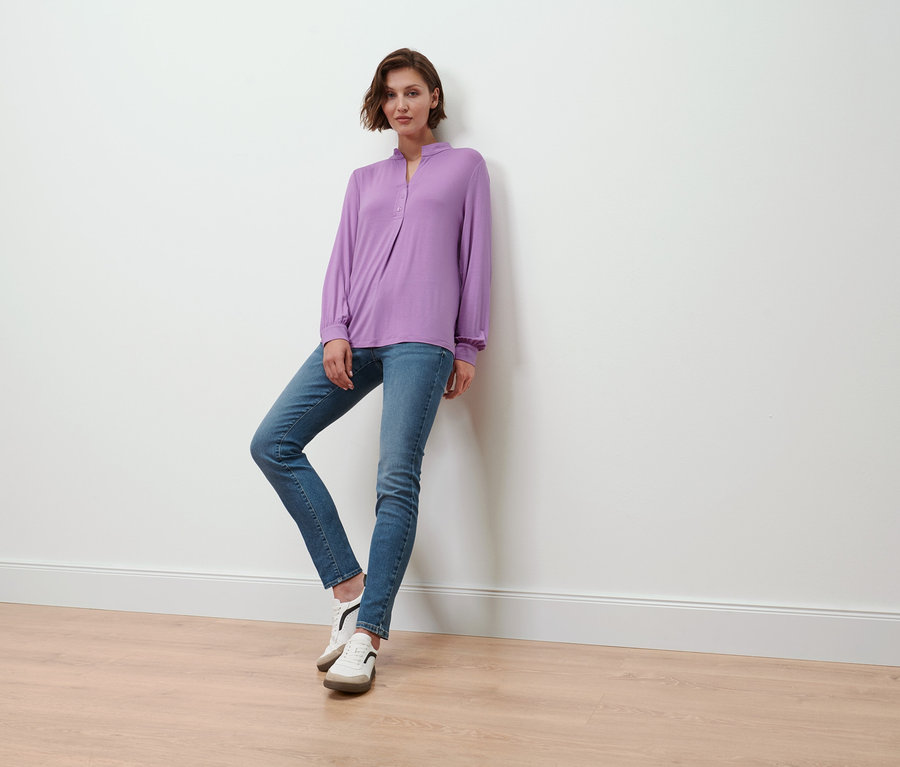 Frau posiert in fliederfarbenem Blusenshirt mit Knopfleiste, blauen Jeans und weißen Schuhen.