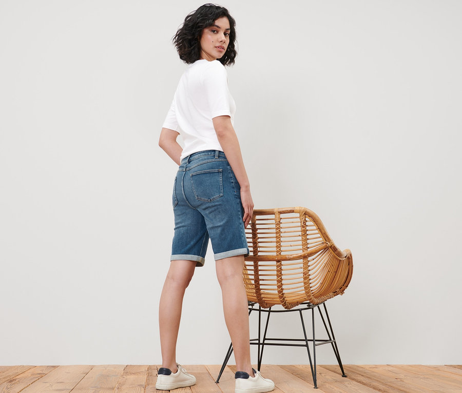 Frau posiert in Bermuda Jeans – Fit »Lea« und weißem T-Shirt neben einem Korbstuhl.