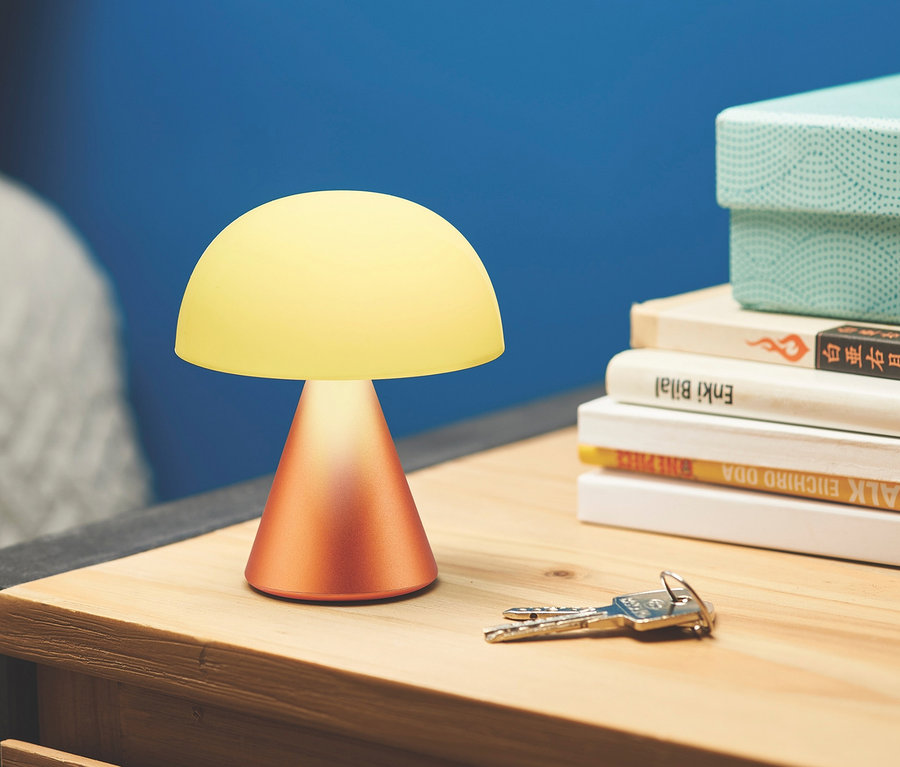 Eine orange LEXON LED-Akku-Tischleuchte »Mina M« steht auf einem hölzernen Nachttisch neben Büchern und Schlüsseln.