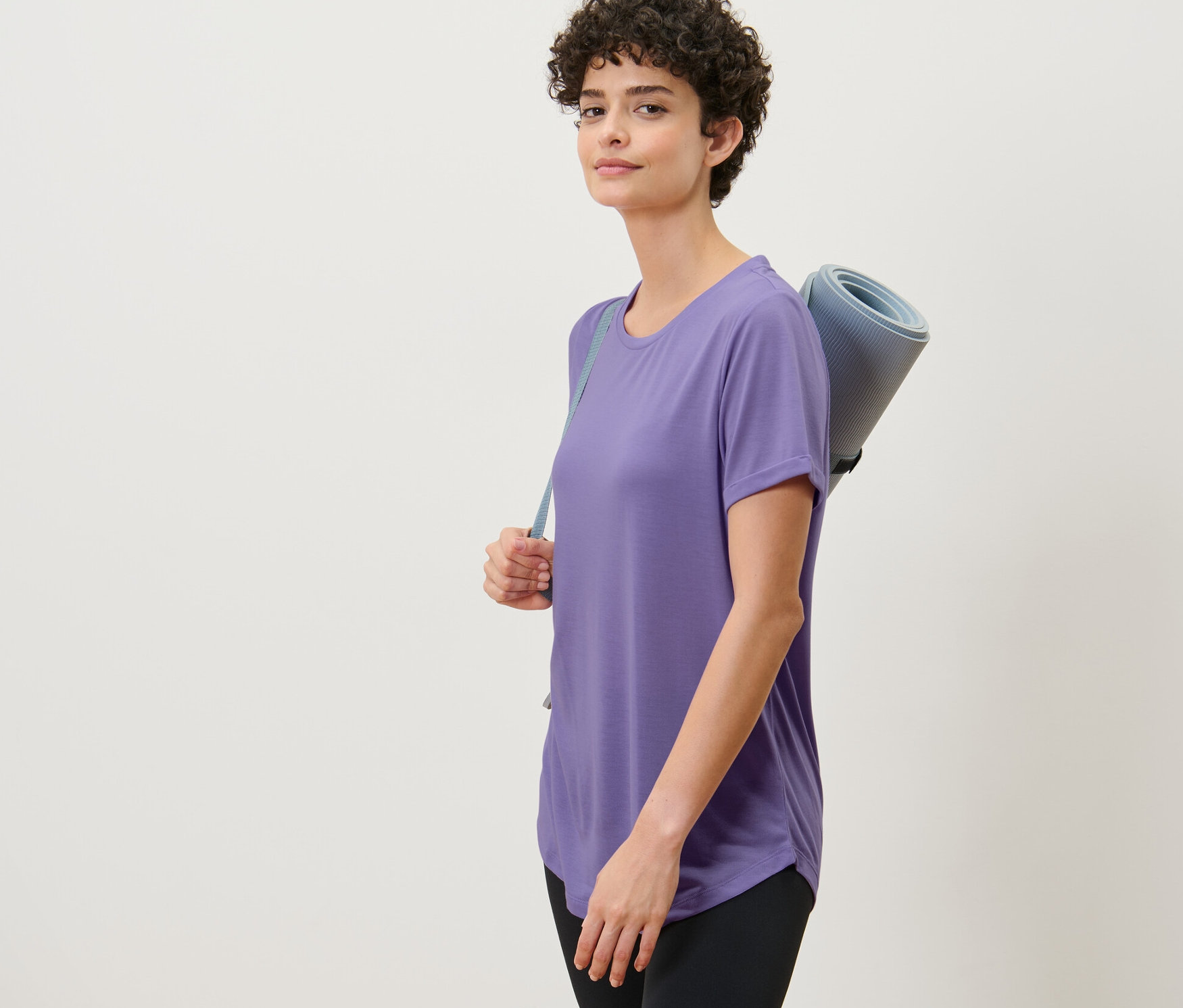 Frau trägt ein lila Longshirt und eine Yogamatte.