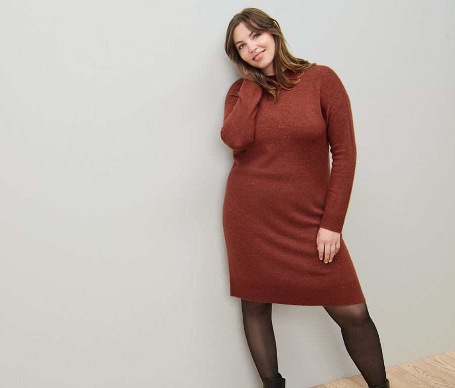 Eine Frau lehnt an einer Wand und trägt ein Strickkleid mit Wollanteil und eine 50-DEN-Feinstrumpfhose in transparenter Optik.