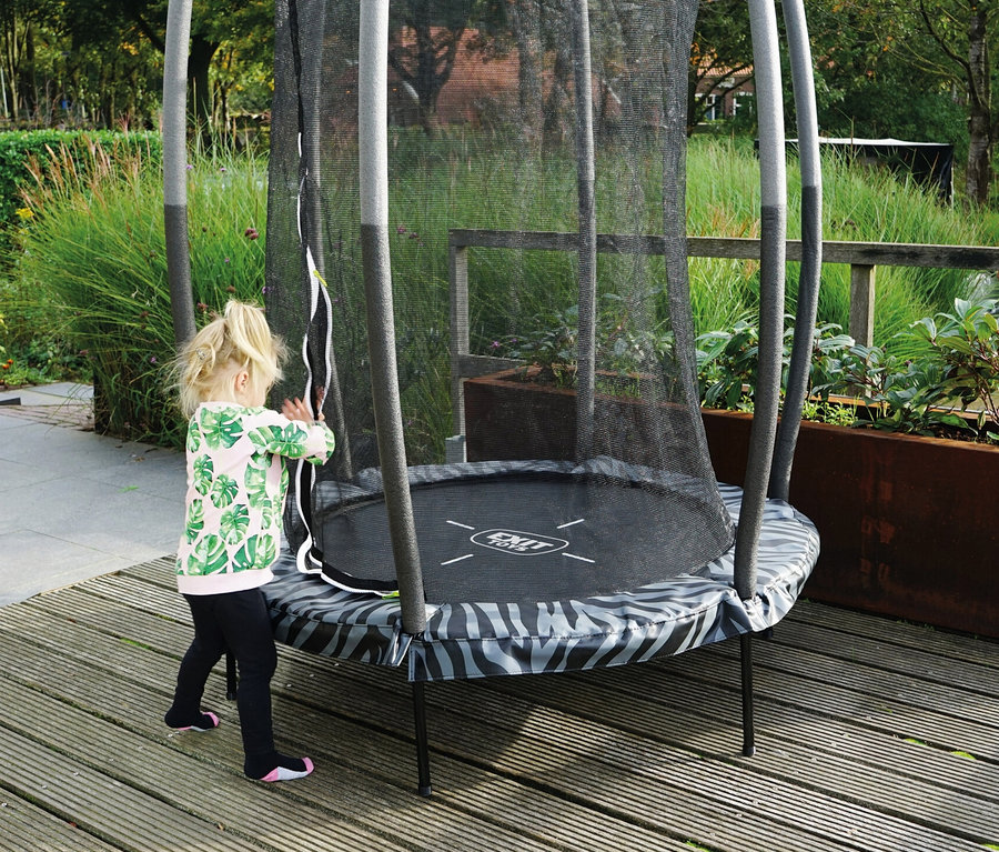 Mädchen berührt das Netz des EXIT Tiggy Junior Trampolins auf einem Holzdeck.