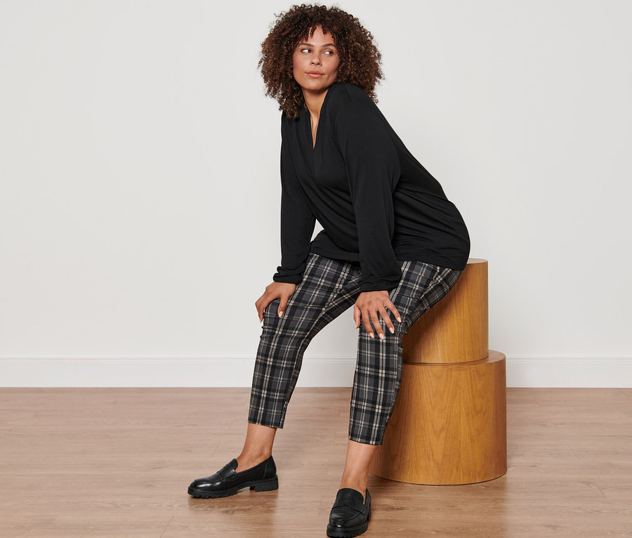 Eine Frau sitzt auf einem Holzhocker und trägt ein schwarzes Langarmshirt mit V-Ausschnitt, eine karierte Stretchhose und Leder-Loafer.