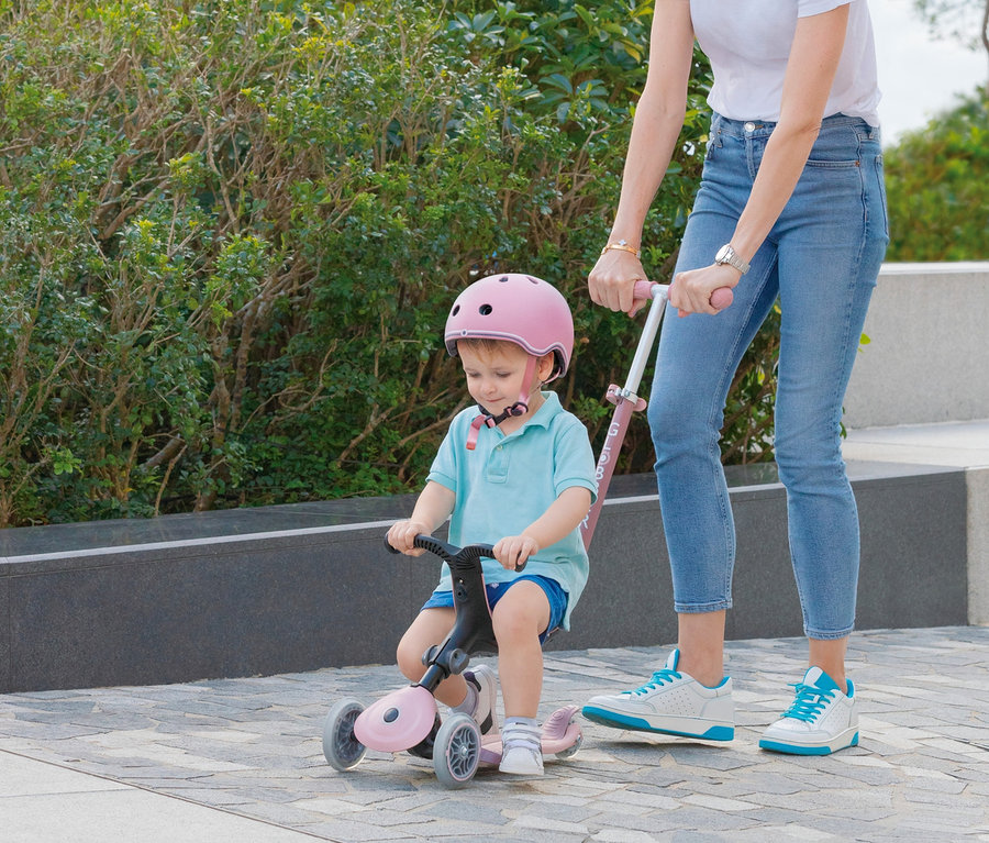Kind mit Helm fährt auf einem himbeerfarbenen GLOBBER Roller »GO-UP Foldable Plus Eco«, während eine Frau es hält.
