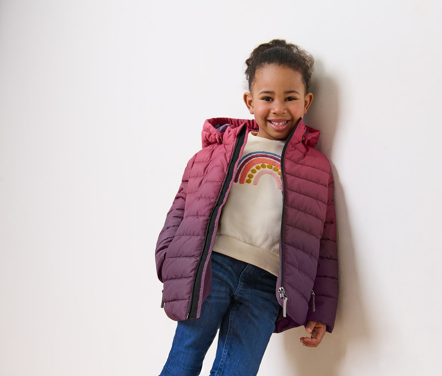 Lächelndes Mädchen posiert in Kinder-Steppjacke mit Farbverlauf.