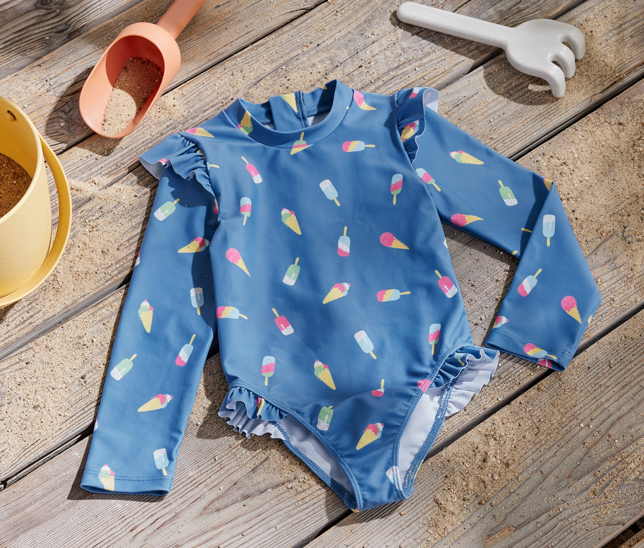 Blauer Kinder-Badeanzug mit Eis-Print und UV-Schutzfaktor 80 liegt neben einem Sandspielzeug auf einem Holzbrett.
