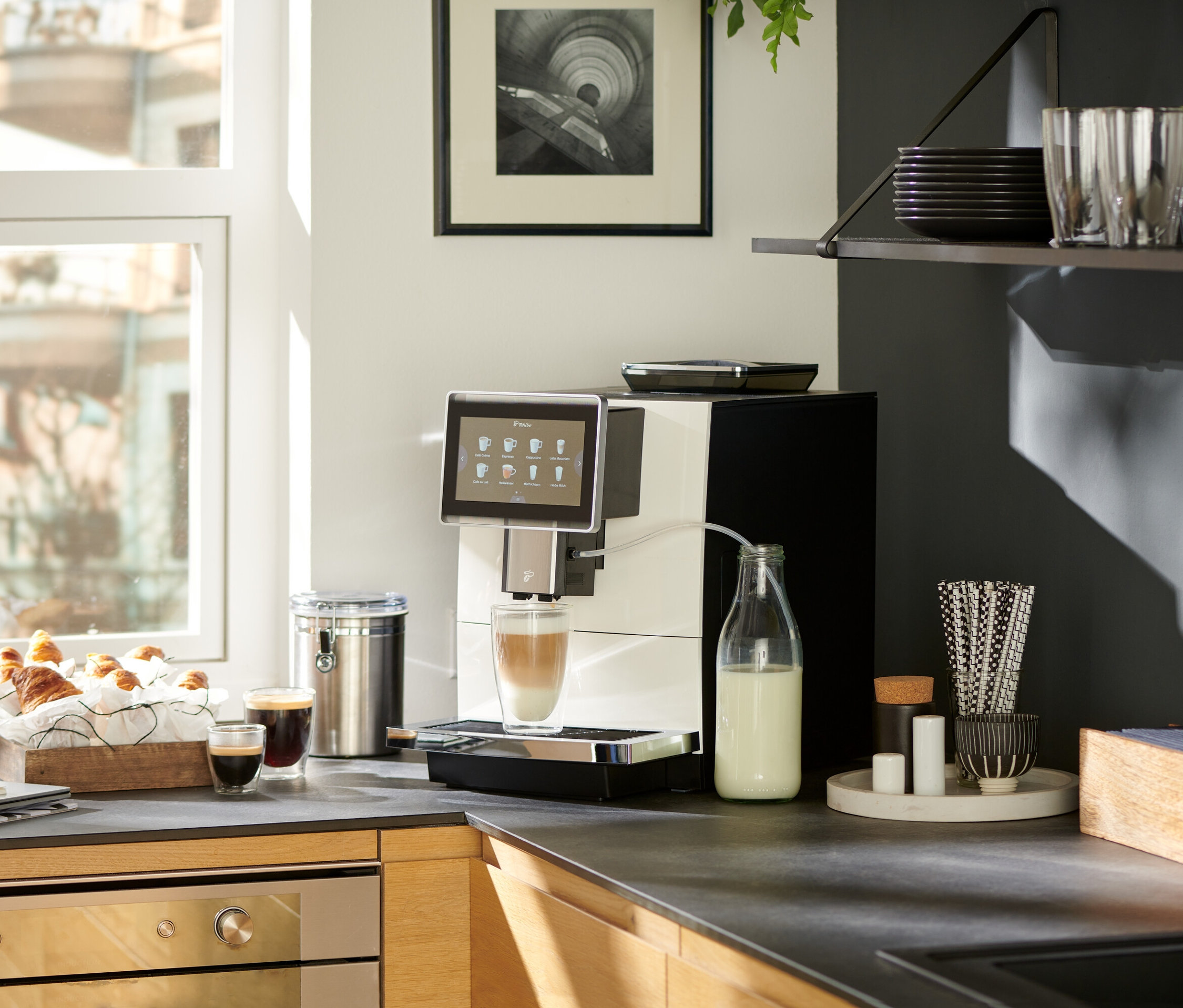 Auf einer Küchentheke steht ein weißer »Tchibo Office« Kaffeevollautomat, der Kaffee in ein Glas zubereitet. Daneben stehen eine Milchflasche, Croissants, Kaffee und ein Notizbuch.