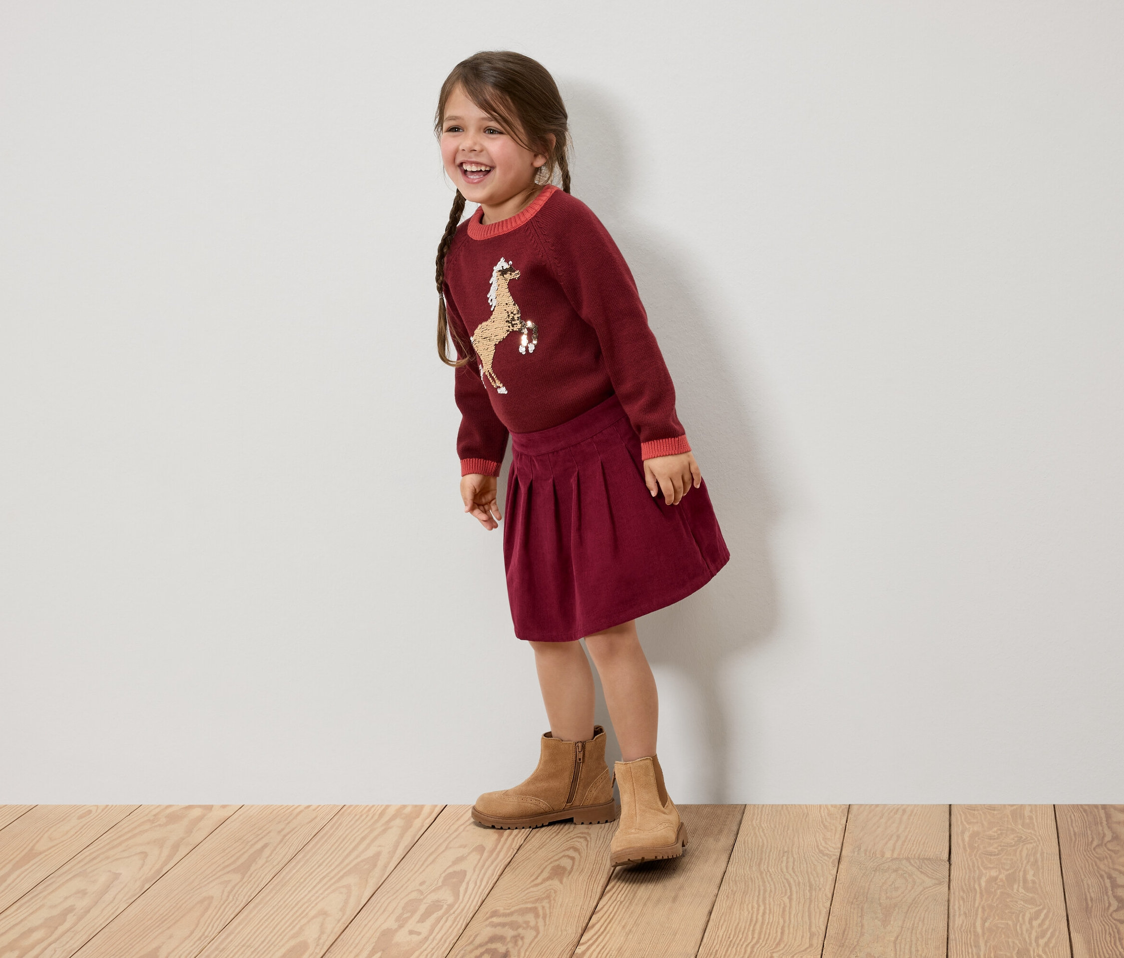 Ein lächelndes Mädchen mit Zöpfen trägt einen bordeauxroten Pullover mit einem Pferd, einen Rock und braune Stiefel.