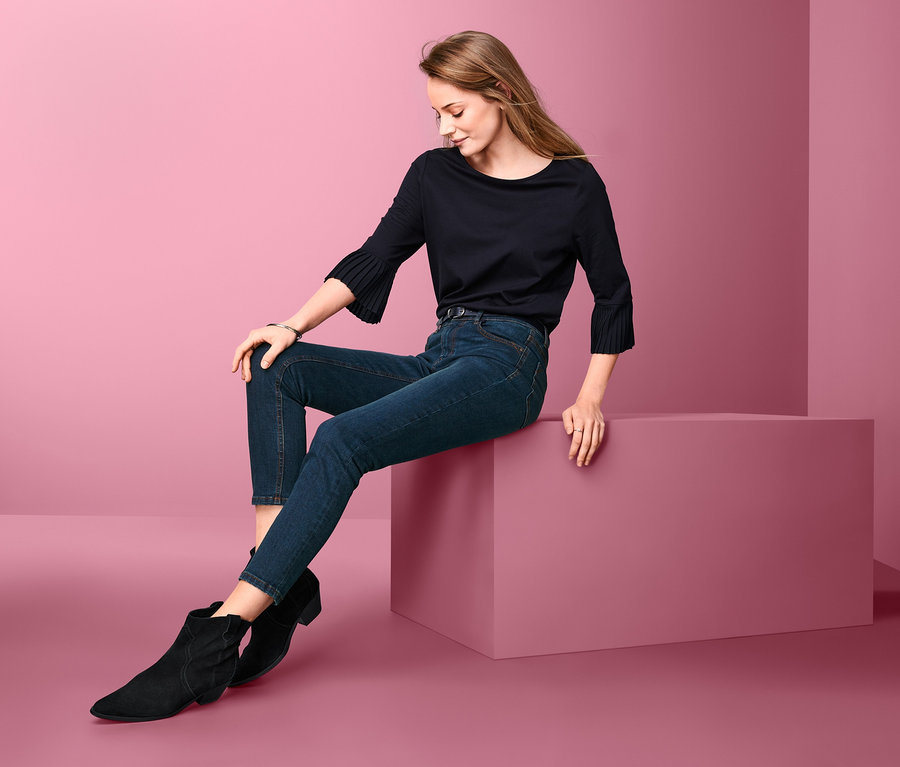 Eine Frau sitzt auf einem Block und trägt eine Slimfit-Jeans, schwarze Stiefel und eine schwarze Bluse.