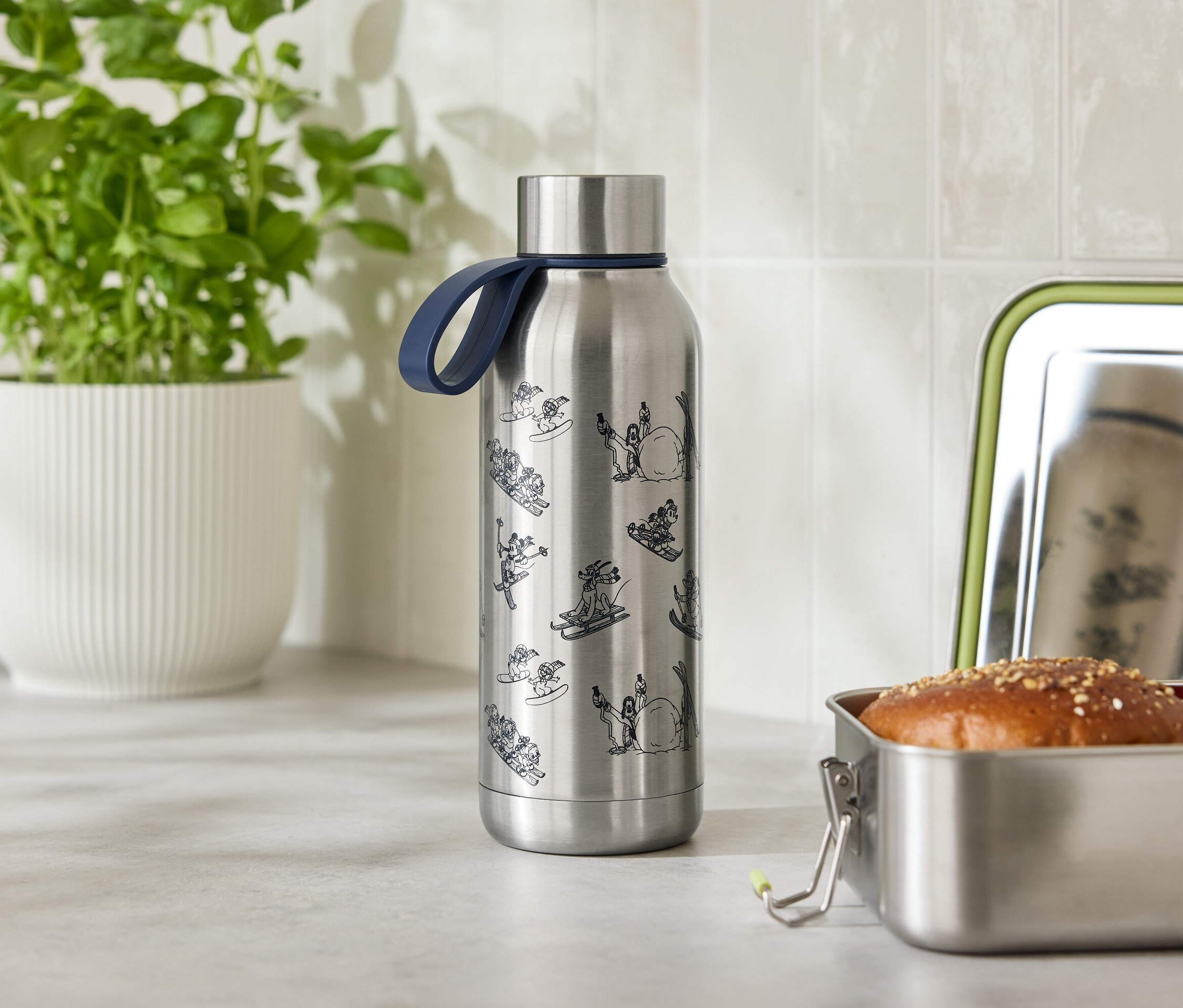 Eine Edelstahlwasserflasche mit blauem Band steht neben einer geöffneten Edelstahl-Lunchbox mit Brötchen und Tomate.