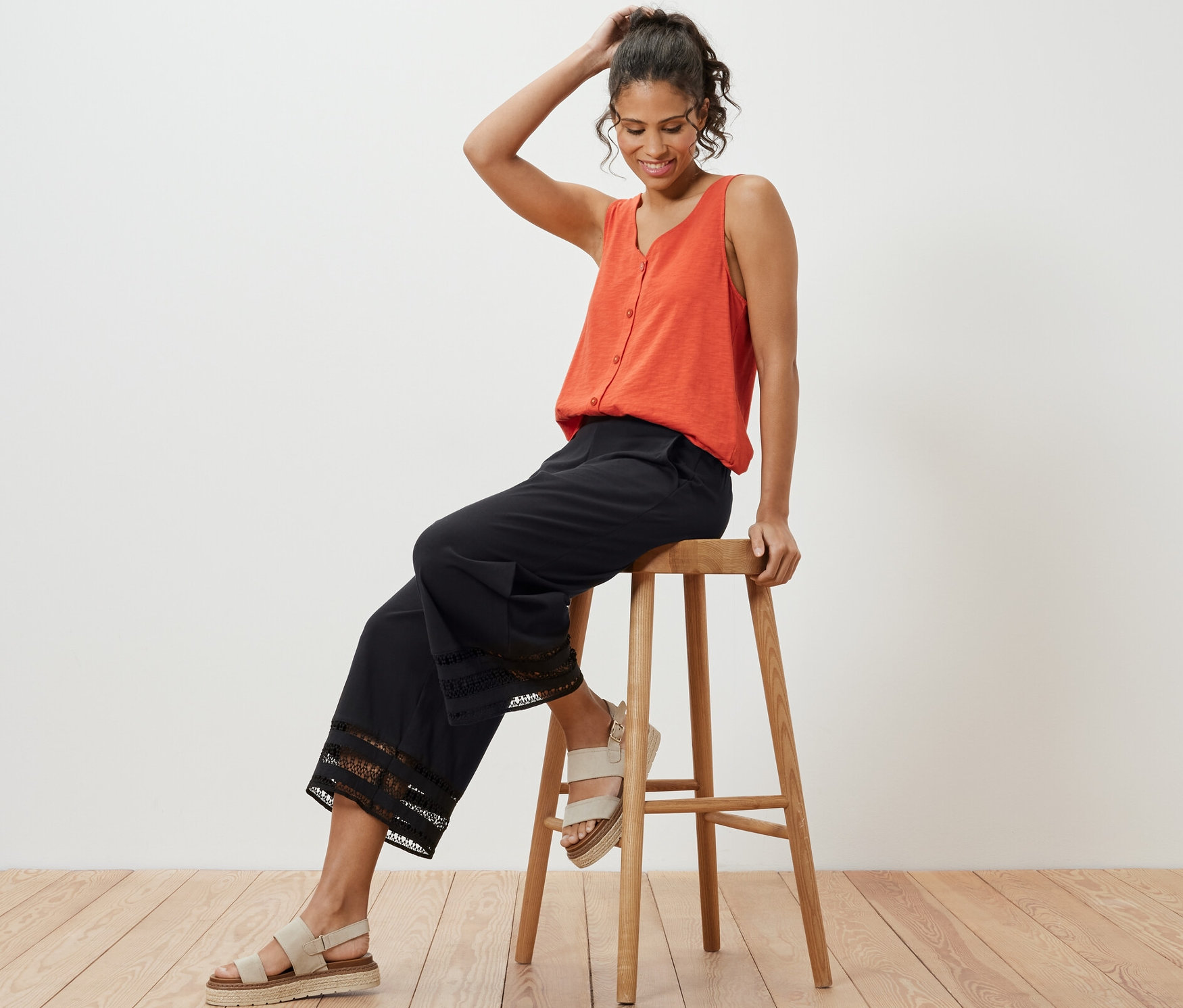 Frau sitzt auf einem Holzschemel, trägt ein orangefarbenes Tanktop und eine schwarze Culotte mit Stickerei.