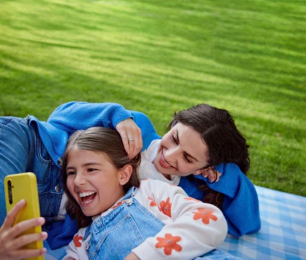 Mutter und Tochter liegen auf einer Decke im Gras. Das Mädchen lacht und hält ein gelbes Telefon.