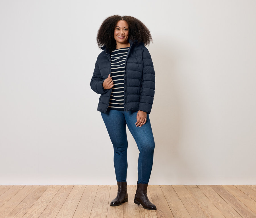 Eine Frau mit lockigem Haar steht und trägt eine blaue Steppjacke, ein gestreiftes T-Shirt, Jeans und Stiefel.