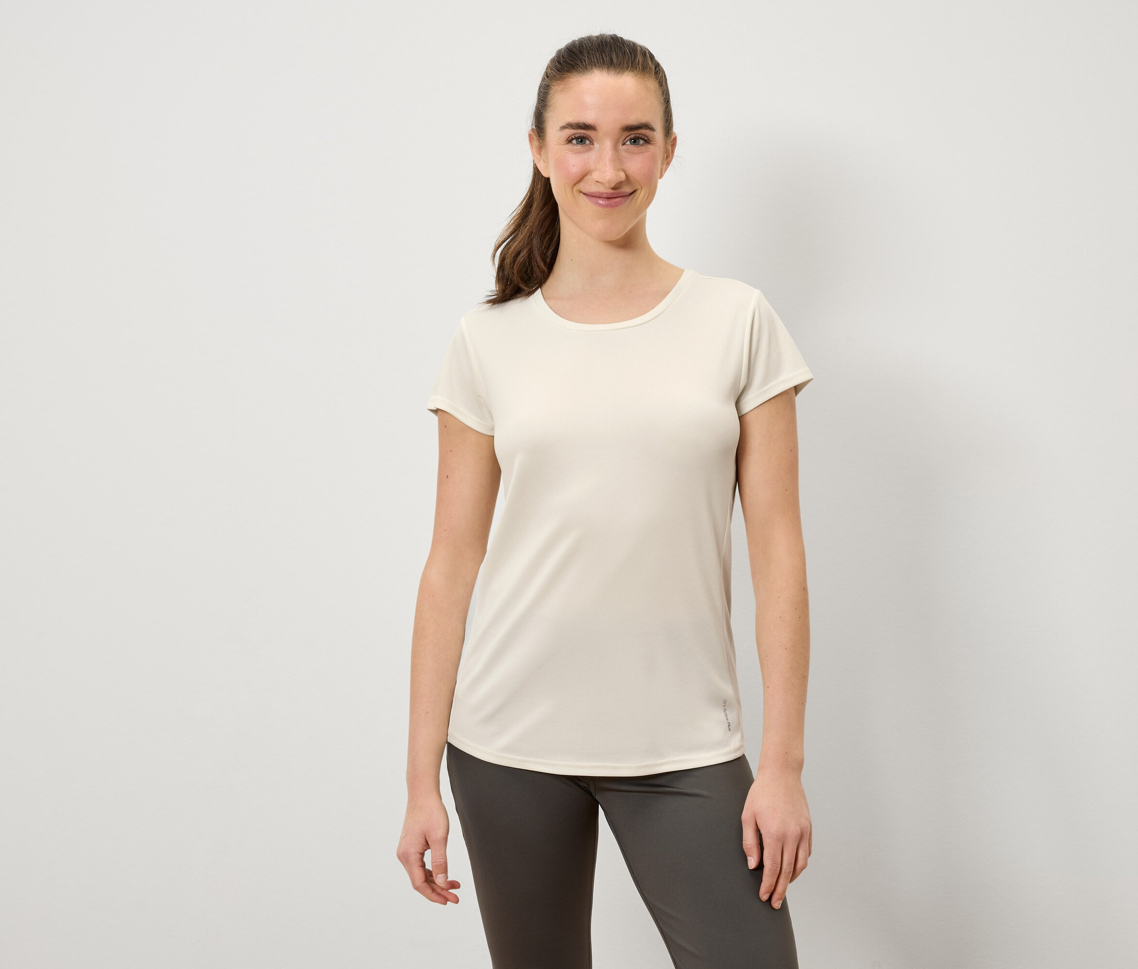 Lächelnde Frau in einem cremefarbenen T-Shirt und grauer Hose posiert vor einem weißen Hintergrund.