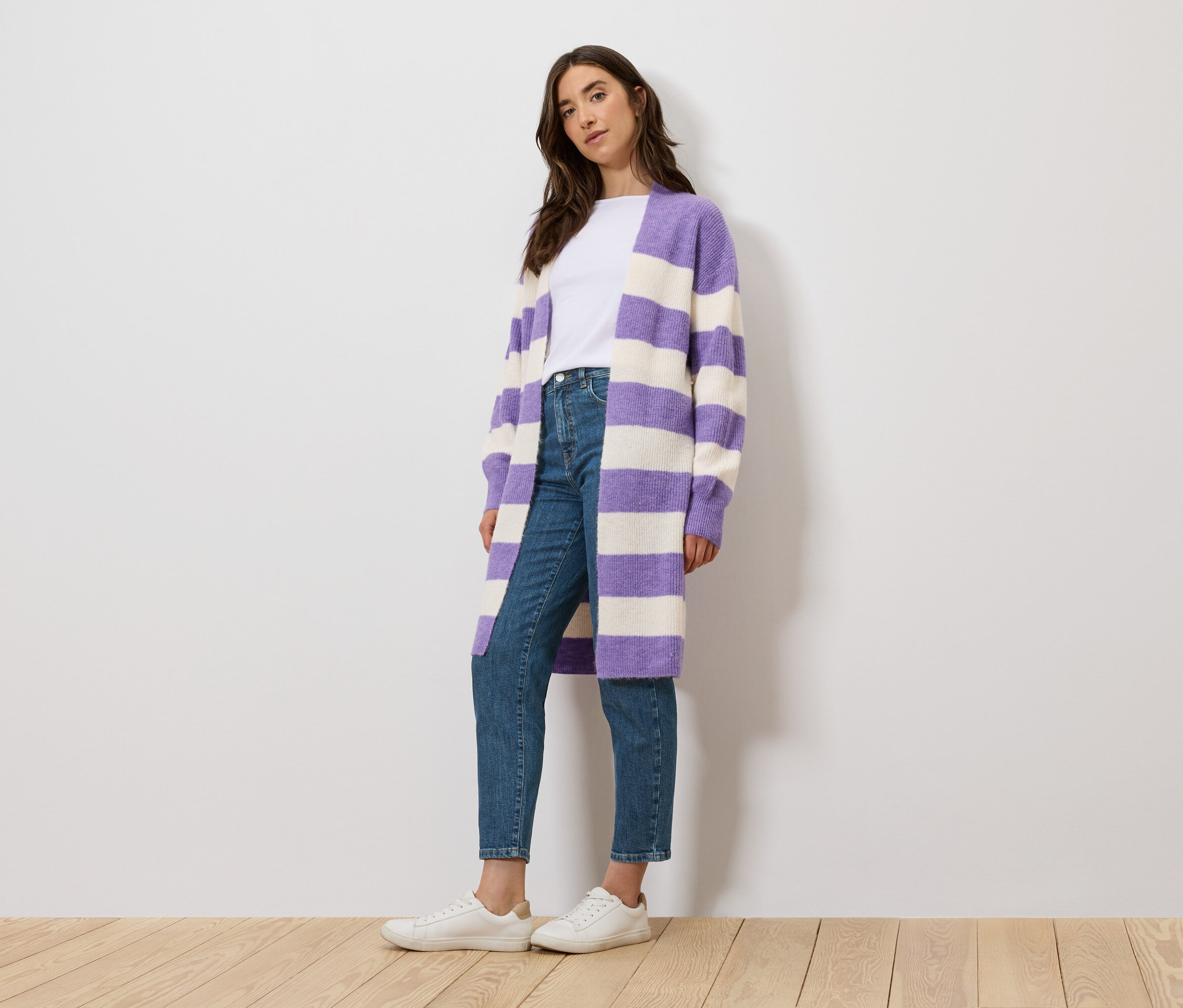 Eine Frau steht und trägt ein weißes T-Shirt, Jeans und einen lila-weiß gestreiften Cardigan.