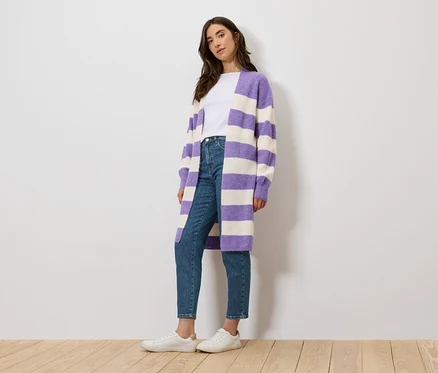 Eine Frau steht und trägt ein weißes T-Shirt, Jeans und einen lila-weiß gestreiften Cardigan.