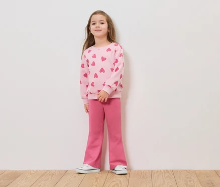 Mädchen steht vor einer weißen Wand in einem rosa Pullover mit Herzen und rosa Hosen.