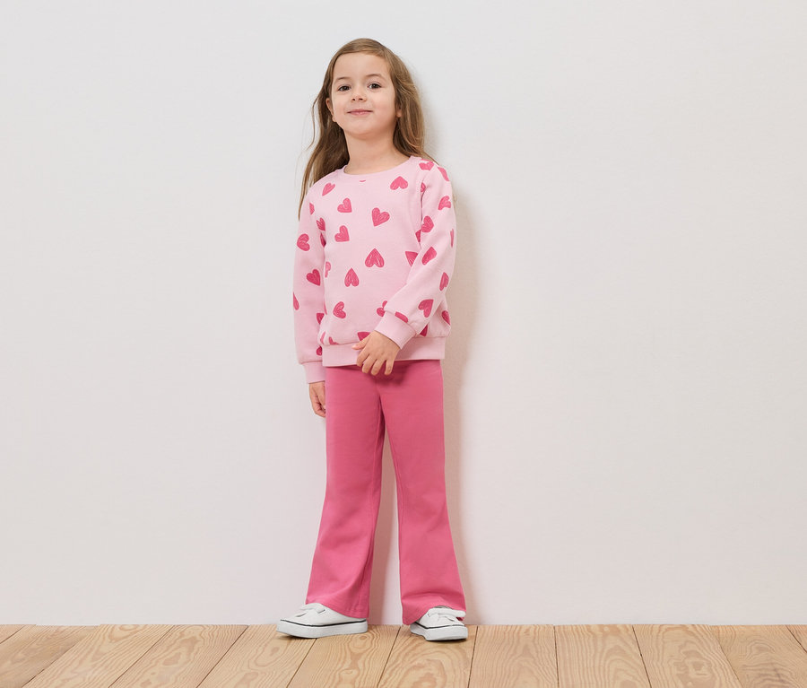 Mädchen steht vor einer weißen Wand in einem rosa Pullover mit Herzen und rosa Hosen.