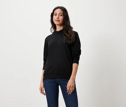 Eine Frau mit langen braunen Haaren steht und trägt einen schwarzen Hoodie und Jeans.