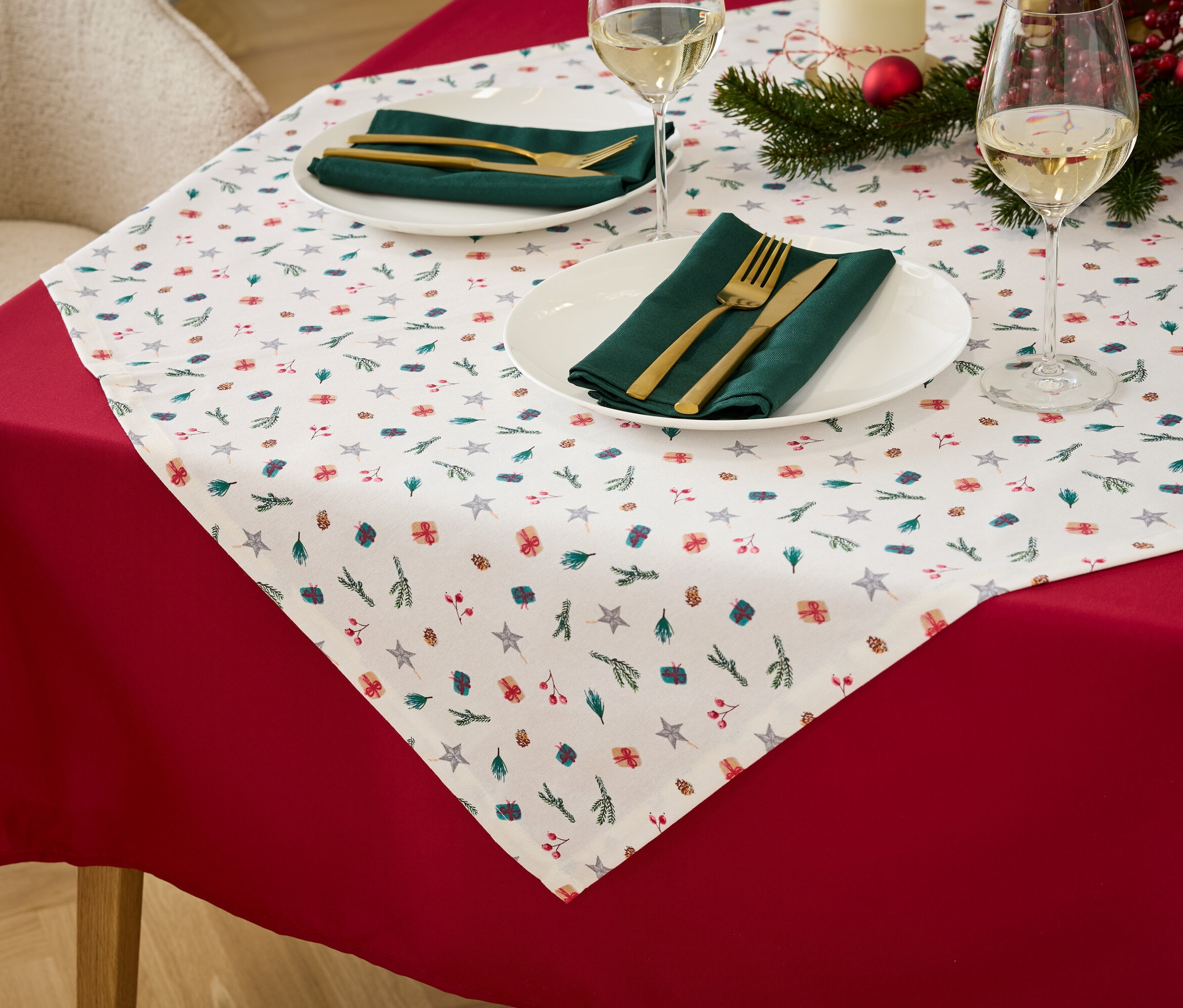 Detailansicht eines Tisches mit roter Tischdecke, weißem Tischläufer mit Weihnachtsmotiv, grünen Stoffservietten und goldenem Besteck.