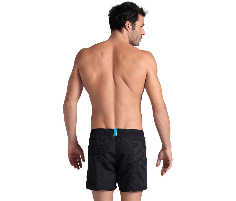 Mann von hinten, oberkörperfrei, trägt schwarze arena Pro_File Beach Shorts mit blauem Detail am Bund, blickt über seine rechte Schulter.