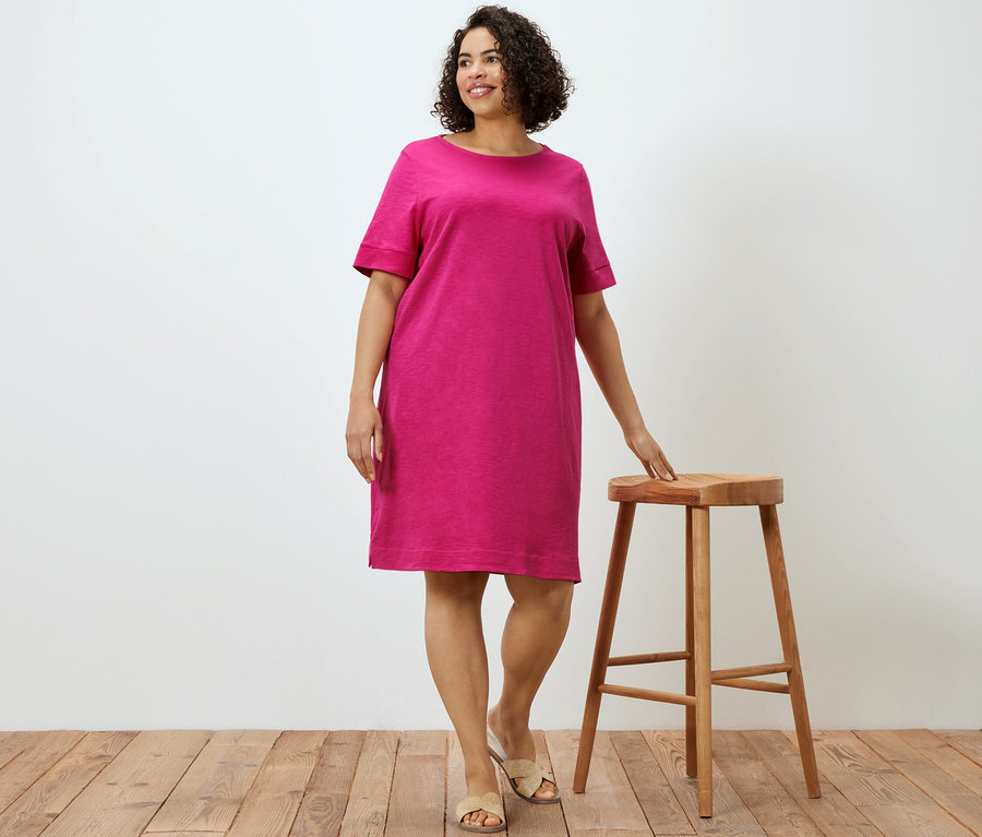 Frau posiert neben einem Holzhocker und trägt ein pinkes Jerseykleid.