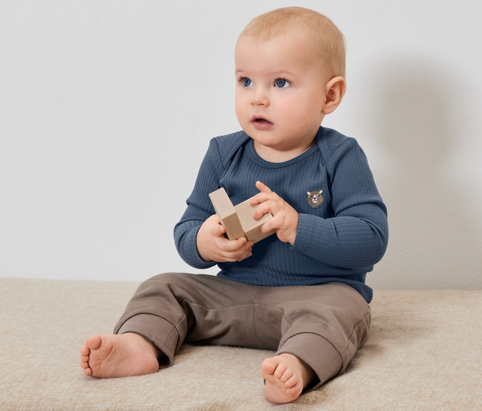 Ein Baby sitzt und spielt mit Holzspielzeug. Es trägt ein blaues geripptes Langarmshirt.