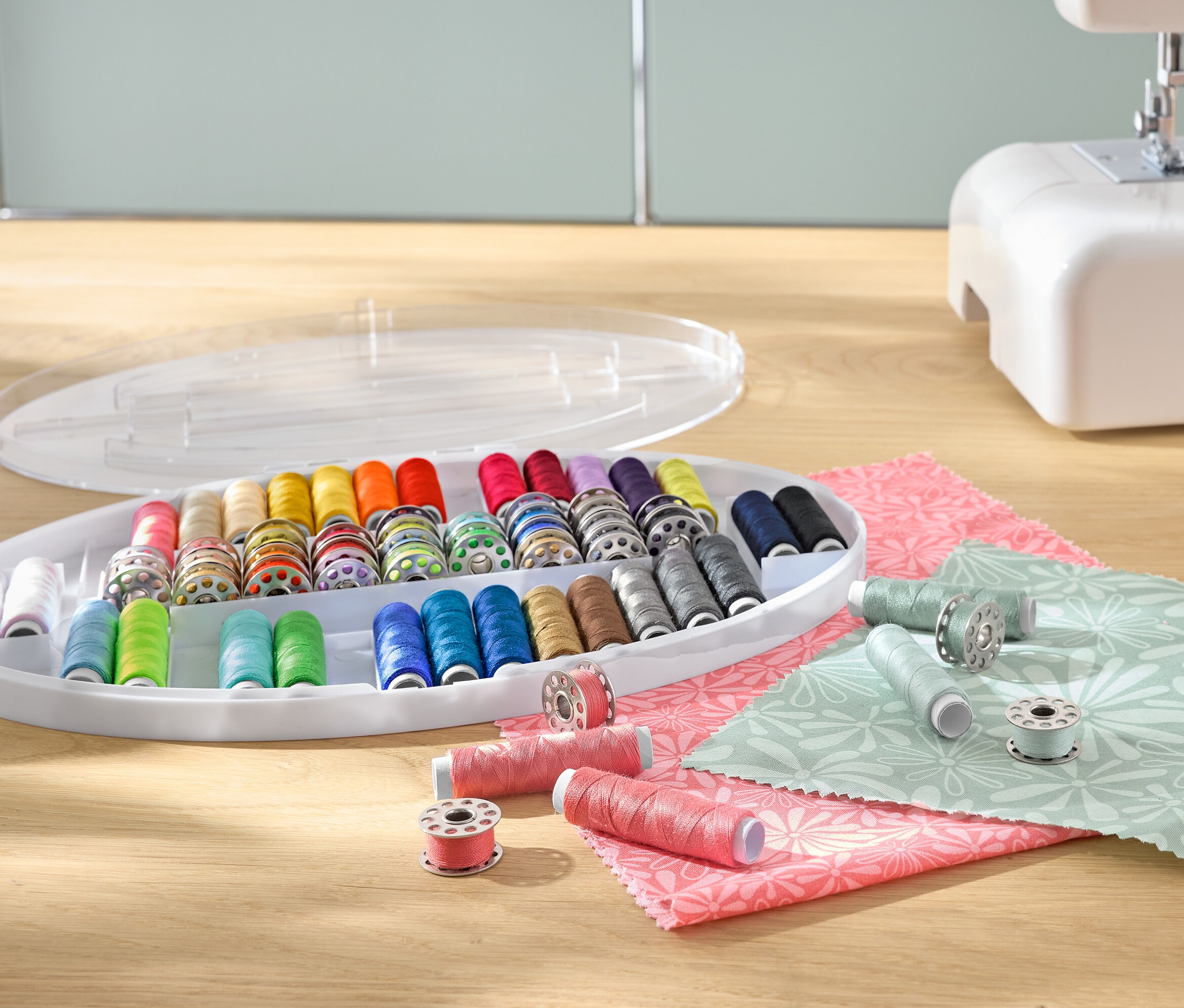 Ein Nähgarn-Set mit bunten Garnspulen in einem Organizer aus Kunststoff, neben einer Nähmaschine und Stoffen.