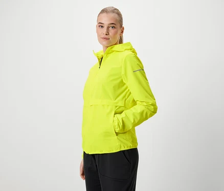 Blonde Frau in neongelber Kapuzenjacke und schwarzen Hosen posiert seitlich.