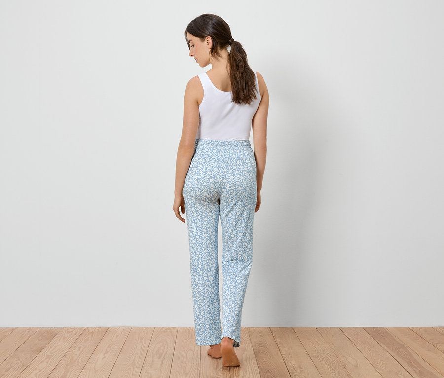 Eine Frau von hinten, gekleidet in ein weißes Tanktop und hellblaue Hosen mit Blumenmuster, steht auf einem Holzboden.